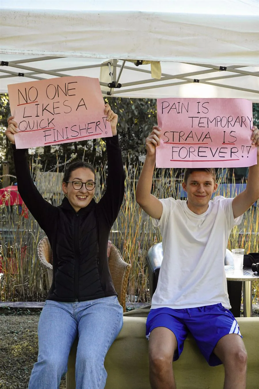 Zwei Fans unter dem Zelt mit Schildern: No one likes a quick finisher, Pain is temporary – Strava is forever
